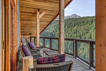 A rustic wooden balcony with lounge chairs and plaid cushions overlooks a dense pine forest and rugged mountains under a clear sky.