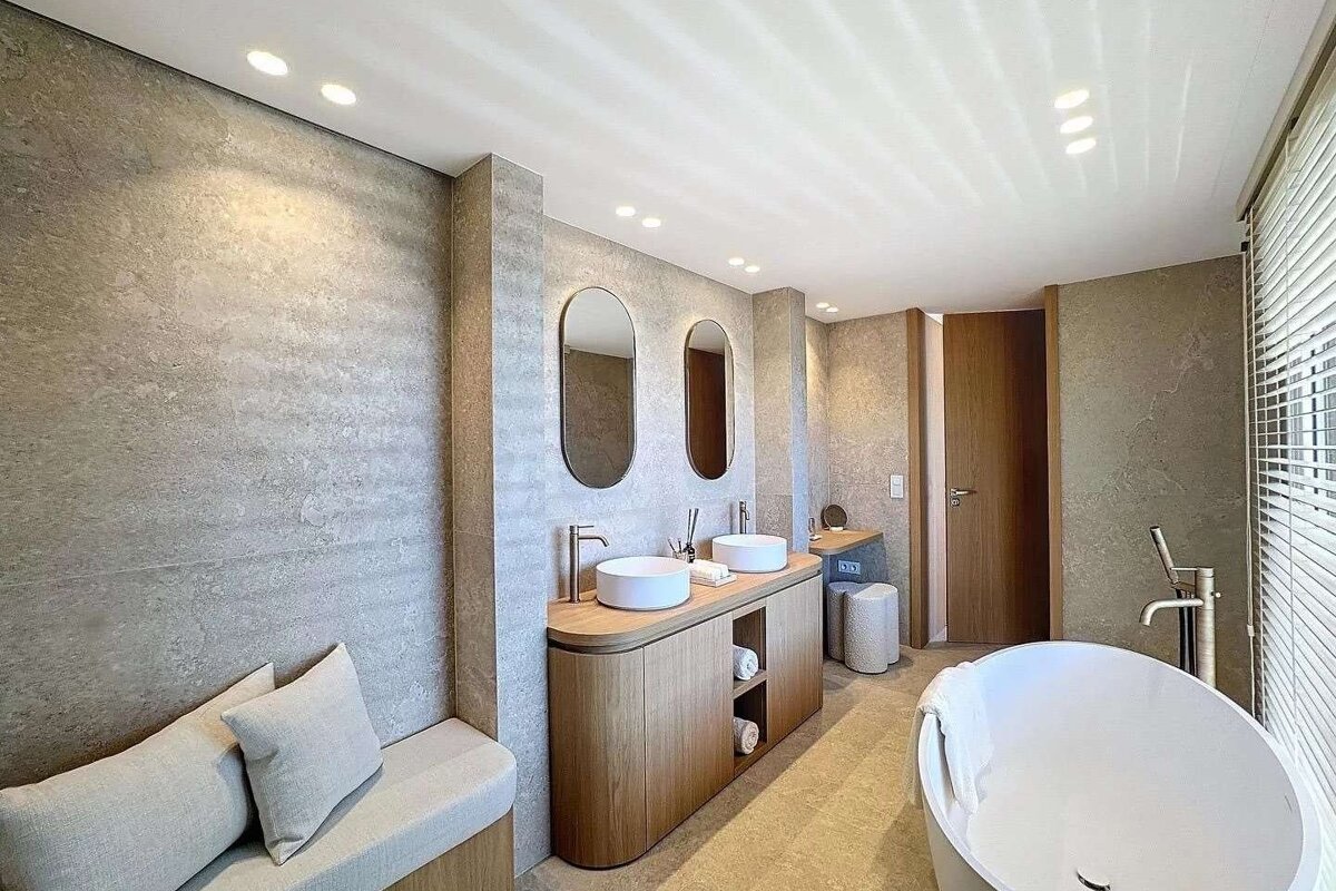 A modern, spacious bathroom with neutral gray walls, a wooden double vanity with dual sinks, a freestanding tub, and a built-in bench.