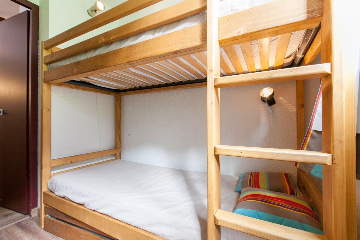 A set of light wood bunk beds with white mattresses. The lower bunk has a colorful striped pillow, and a reading light is mounted on the wall.