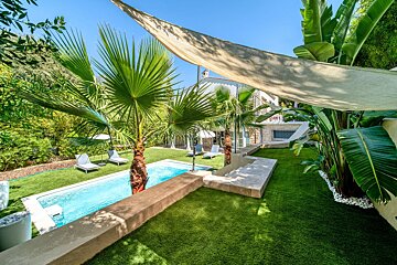 A tropical pool area with lush green lawn, palm trees, lounge chairs, and a house, all bathed in sunlight under a large shade sail.