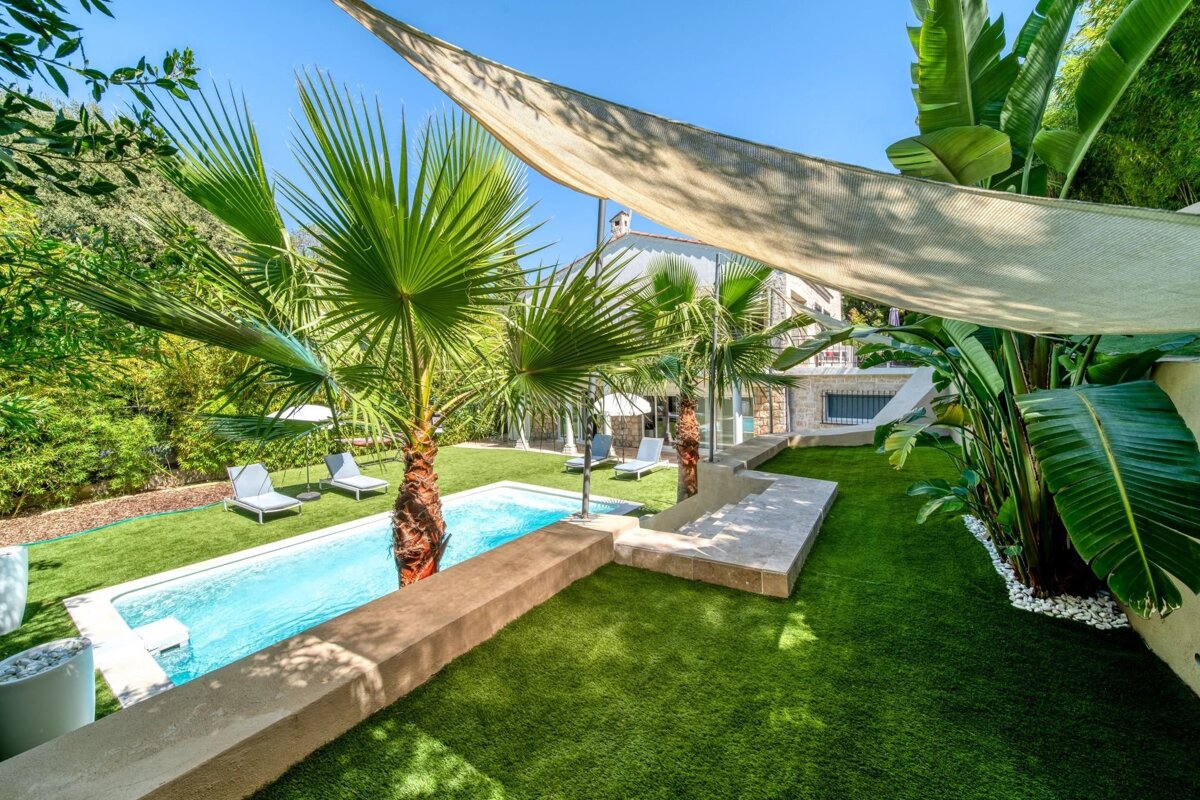 A backyard swimming pool surrounded by a green lawn, palm trees, banana plants, lounge chairs, and a shade sail under a clear blue sky.