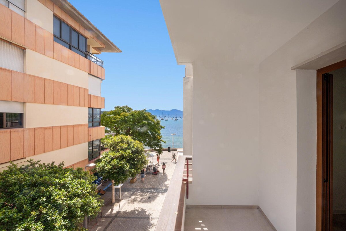A sunny balcony overlooks a coastal town with a building, green trees, a street, a sea full of boats, and distant mountains.