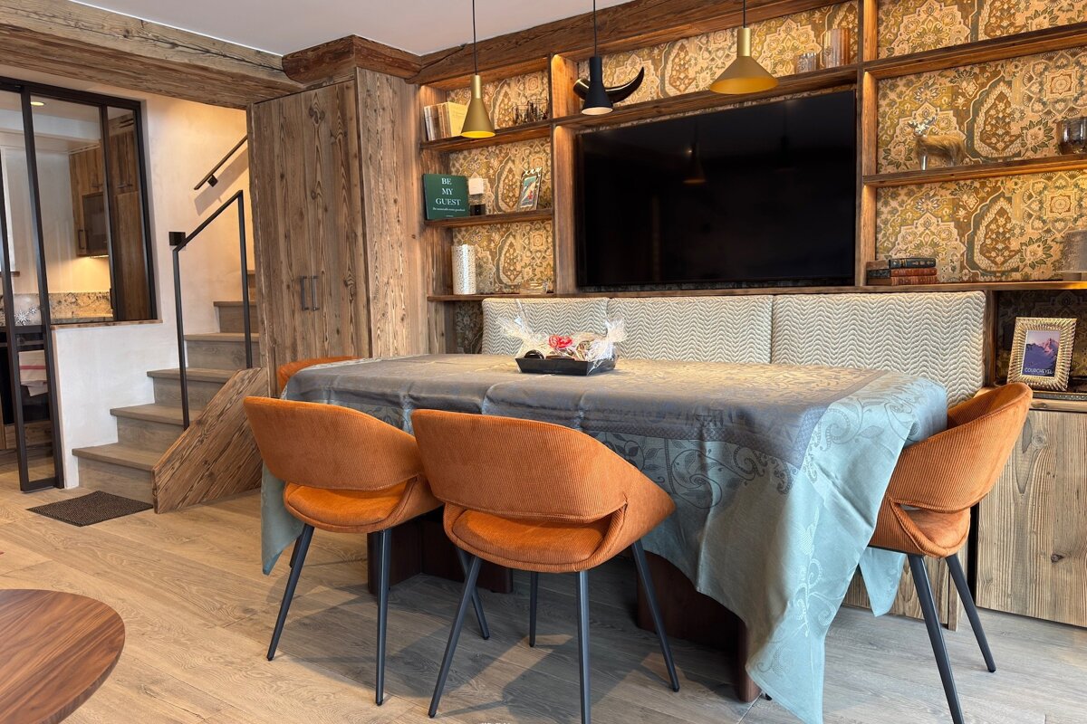 Rustic dining area with a wood-paneled wall, built-in TV, a table covered in a green cloth, and three bright orange chairs.