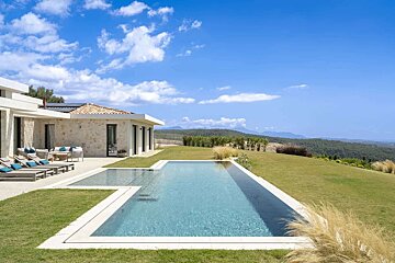 A luxurious stone villa with a modern L-shaped pool overlooks rolling green hills under a bright blue, cloudy sky. Outdoor lounge seating invites relaxation.