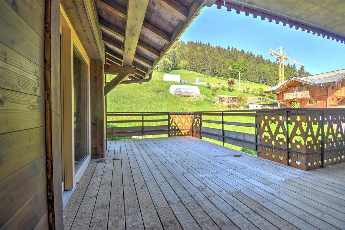 A spacious wooden balcony with an ornate railing overlooks a green mountain with a forest, a construction crane, and other buildings under a clear sky.