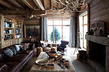 A rustic wooden chalet living room with leather furniture, an antler chandelier, a stone fireplace, and a large window showing a pine tree.
