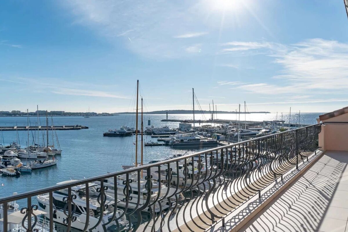 Sunny balcony view of a bustling harbor filled with numerous yachts and sailboats under a bright blue sky, with an ornate railing in the foreground.