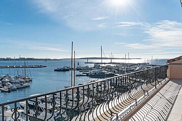 Sunny balcony view of a bustling harbor filled with numerous yachts and sailboats under a bright blue sky, with an ornate railing in the foreground.