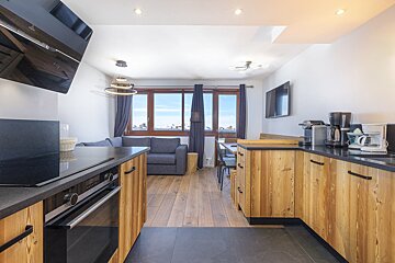 A modern wooden kitchen opening up to a living area with a grey couch and large windows showcasing snowy mountains.