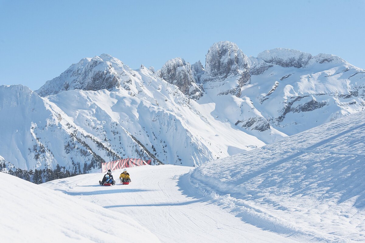 Two people sledding down a sunny, snowy mountain slope, with grand snow-capped peaks dominating the background.