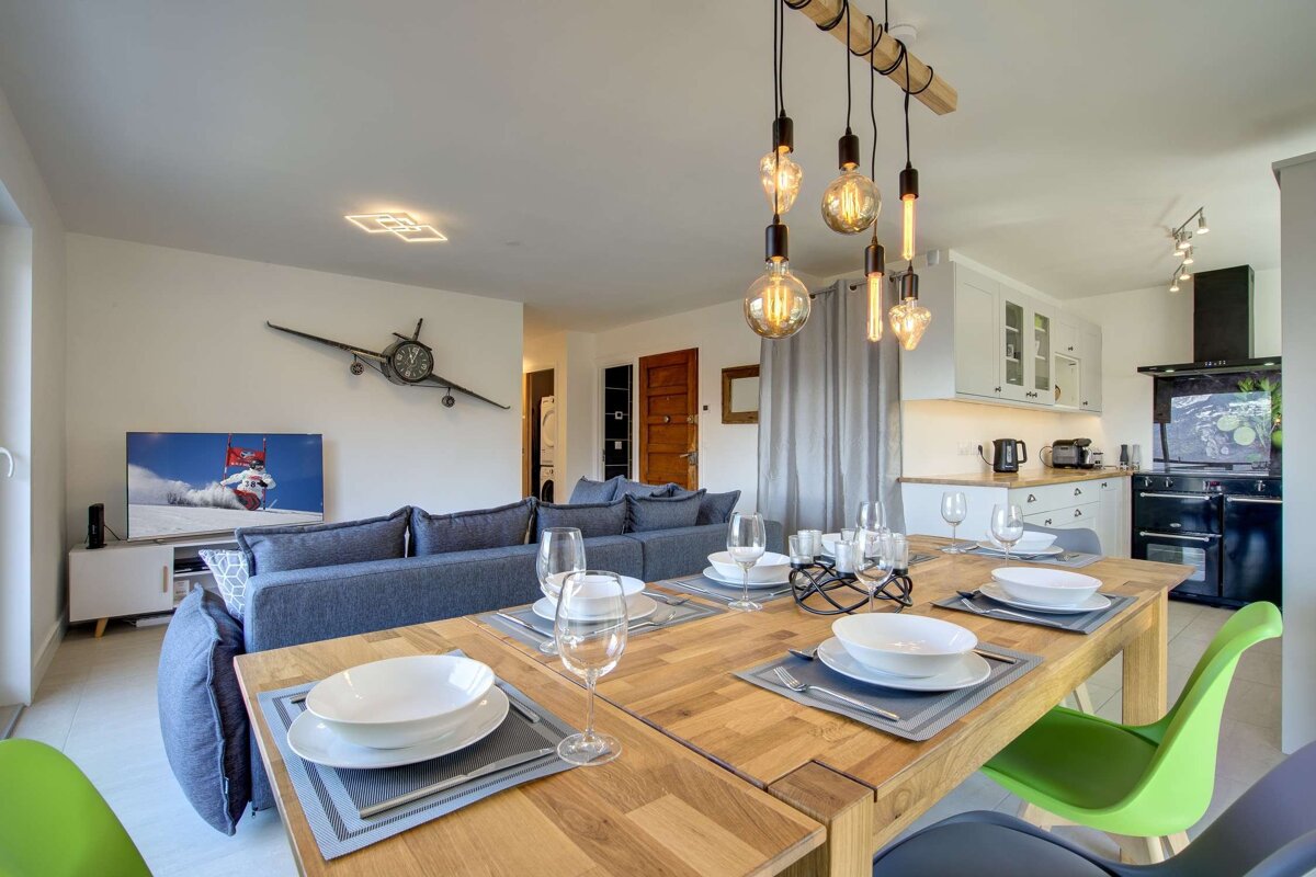 Modern open-concept living space featuring a wooden dining table with place settings, unique pendant lights, a blue sofa, and a kitchen.
