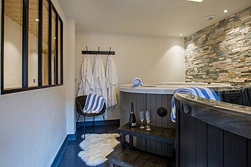 Luxurious indoor spa featuring a wooden hot tub, steps with champagne, white bathrobes, and a stone accent wall.