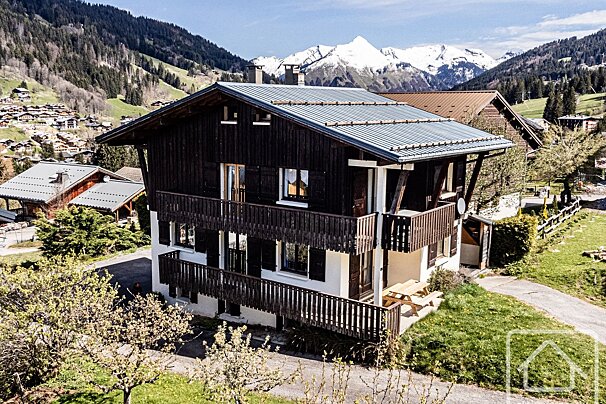 A beautiful alpine chalet with dark wood balconies and white lower level, nestled in a sunny mountain village with snow-capped peaks.