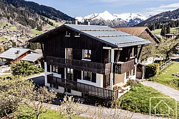 A beautiful alpine chalet with dark wood balconies and white lower level, nestled in a sunny mountain village with snow-capped peaks.