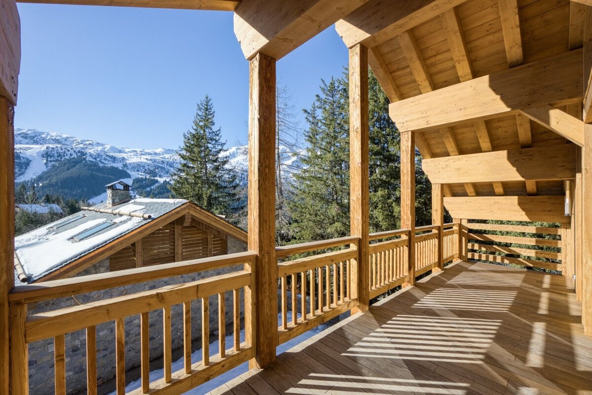 A sunlit wooden chalet balcony with mountain views, pine trees, and snow-covered rooftops on a clear day.