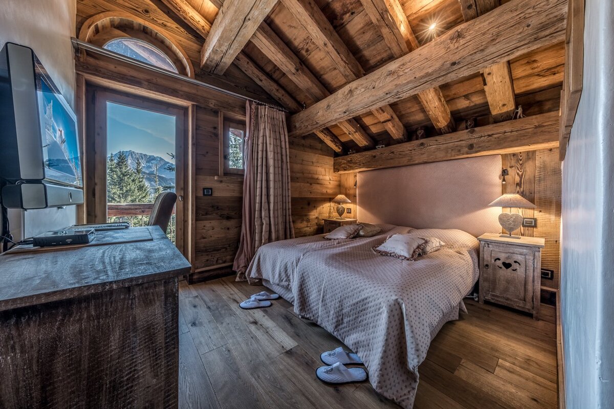Cozy, rustic wooden bedroom with a mountain view balcony, beamed ceilings, and a large bed, featuring a desk and TV.