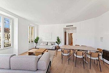 A bright, modern open-plan living and dining room features white walls, light wood floors, grey sofas, potted plants, and a large wooden dining table.