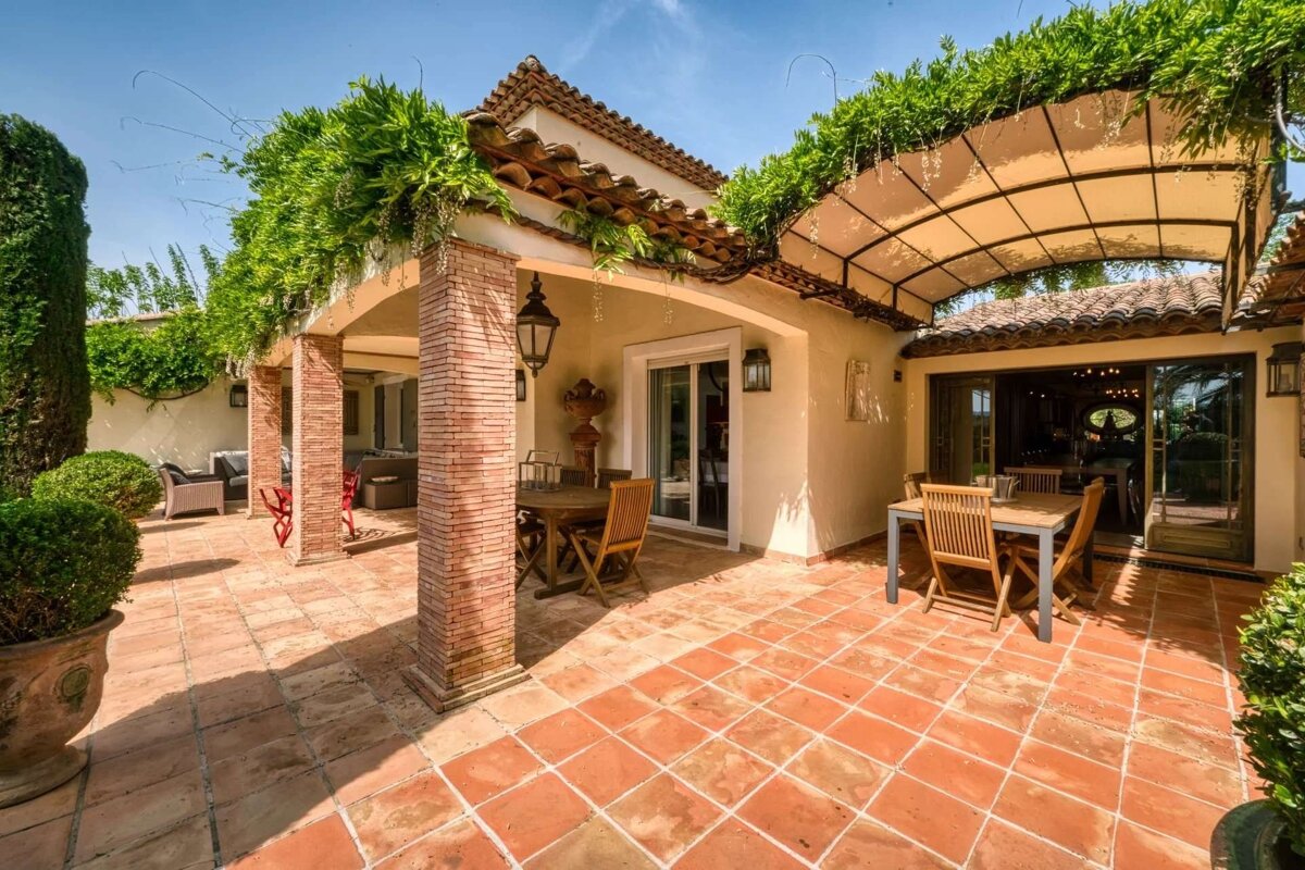 A sunny, terracotta-tiled patio with lush green vines draping over pergolas and an awning. Outdoor dining sets and a Mediterranean-style building are present.