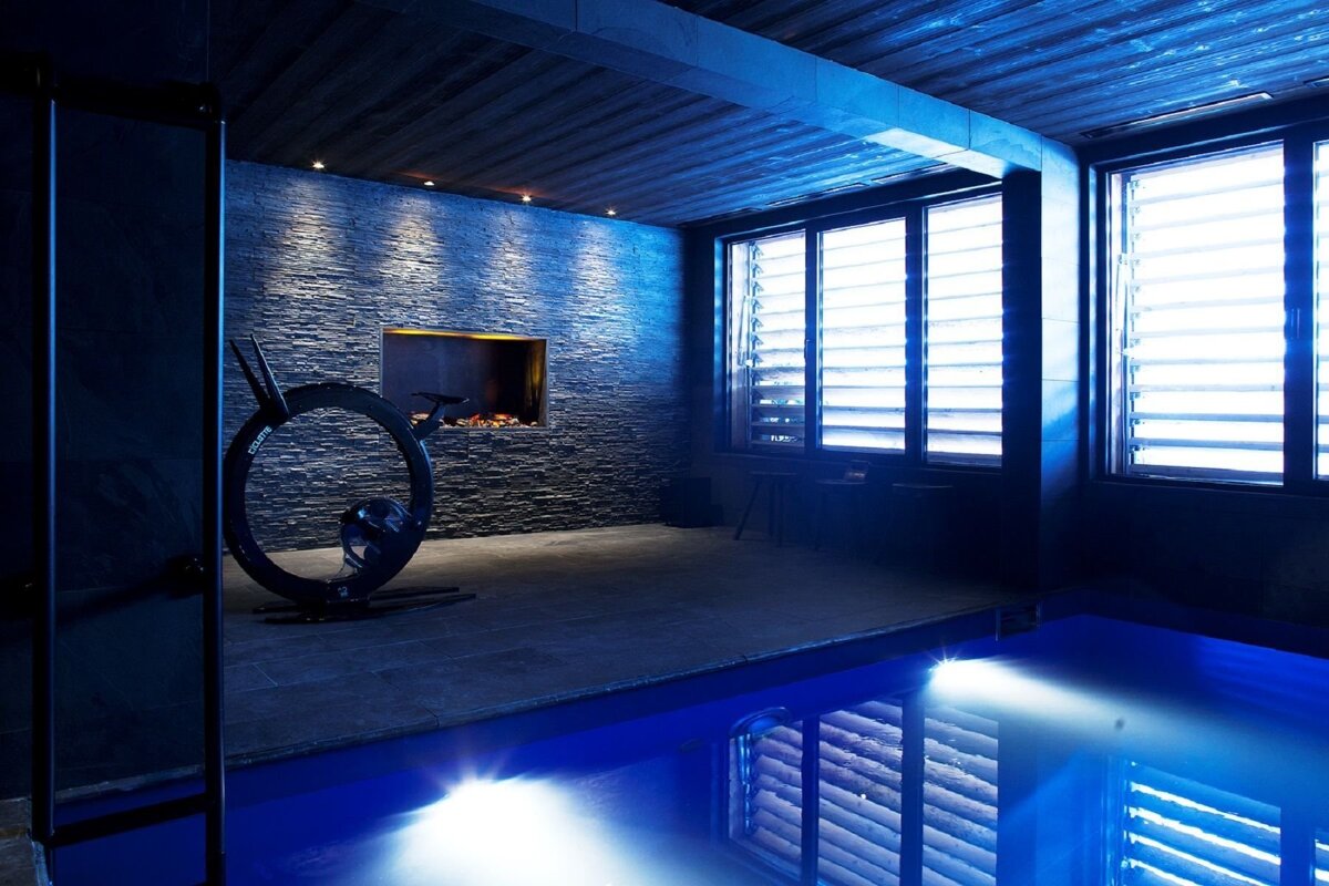 A chic, dimly lit room featuring an indoor pool, a modern exercise bike, a fireplace set in a stone wall, and large louvered windows.