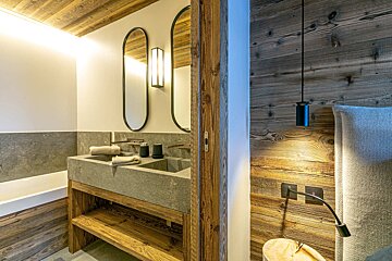 A modern rustic bathroom features a stone double sink on a wooden vanity, with wood-paneled walls extending into a cozy bedroom.