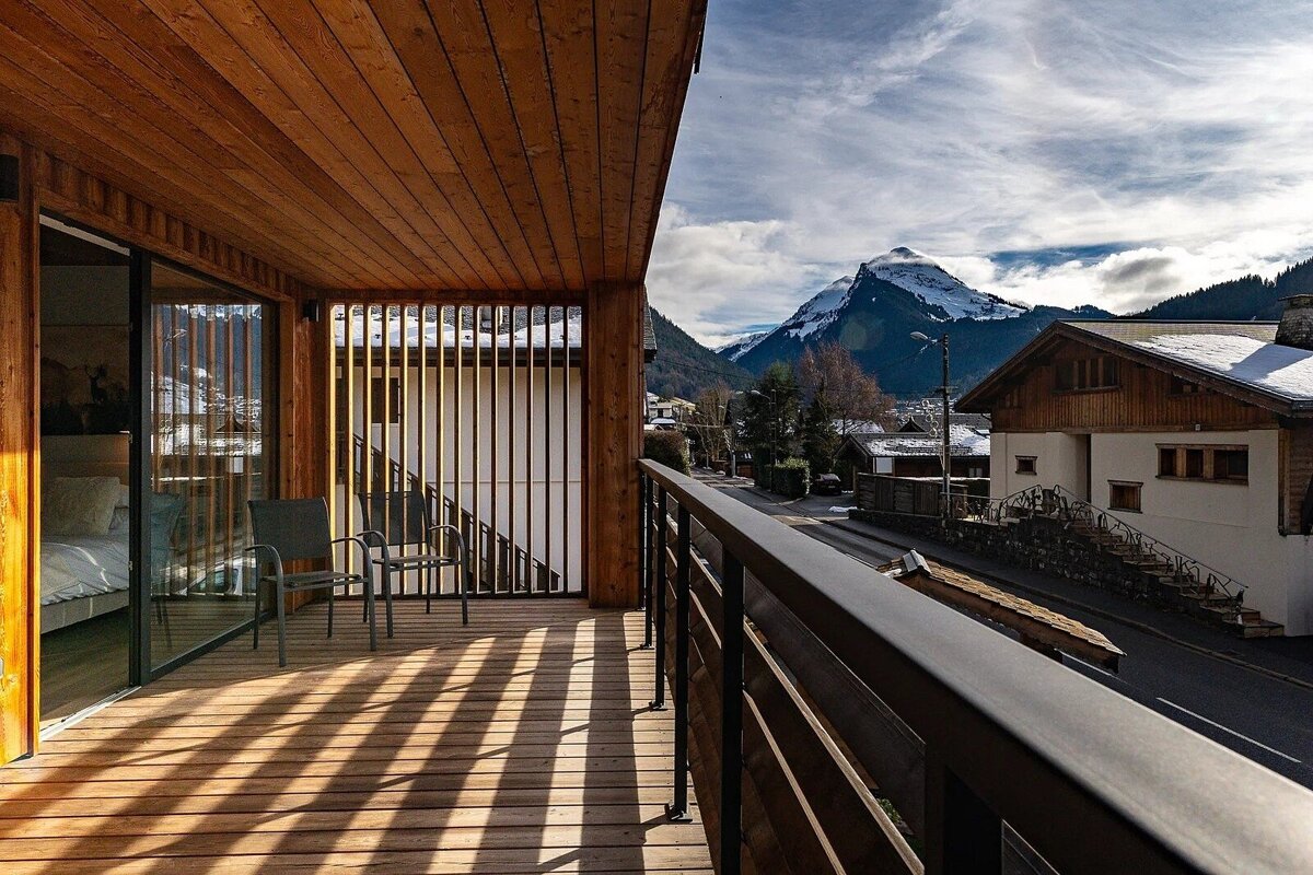 A sunlit wooden balcony with chairs offers a stunning view of a snow-capped mountain and a village below, under a partly cloudy sky.