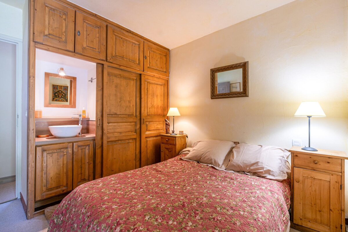 Cozy bedroom featuring a bed with a floral duvet, rustic wooden cabinets, bedside lamps, and a glimpse of an ensuite sink.
