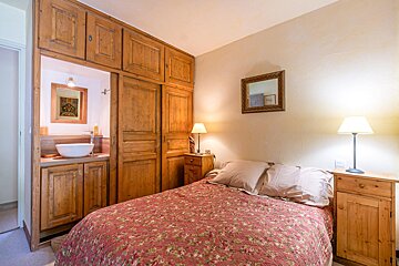 Cozy bedroom featuring a bed with a floral duvet, rustic wooden cabinets, bedside lamps, and a glimpse of an ensuite sink.