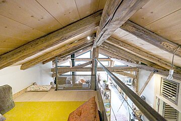 Rustic attic loft with exposed wooden beams, a bright yellow area, triangular windows, and a railing overlooking the lower floor.