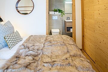 Cozy bedroom with a faux fur blanket on the bed, a round wall mirror, and an open doorway showing a modern bathroom.