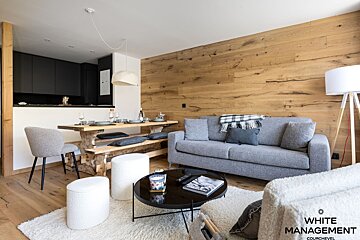 A modern open-plan living and dining area with a rustic touch. Features wood walls and floors, a grey sofa, black kitchen, and a wooden picnic-style dining table.