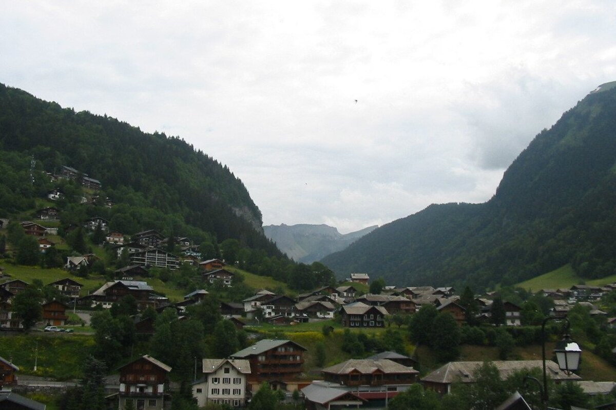 A charming mountain village fills a deep valley, flanked by steep, forested slopes under an overcast sky, with distant peaks visible.
