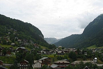 A quaint village nestled in a lush green valley, surrounded by steep forested mountains under a cloudy sky.