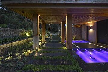 A sleek outdoor path at dusk leads under a wooden canopy, flanked by illuminated landscaping and a vibrant purple-lit water feature.