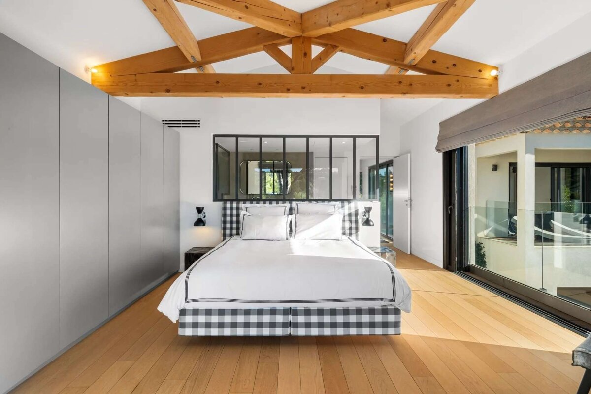 A modern bedroom features exposed wooden beams, a black and white gingham bed, light wood floors, and a glass partition, with a sliding door leading to a balcony.