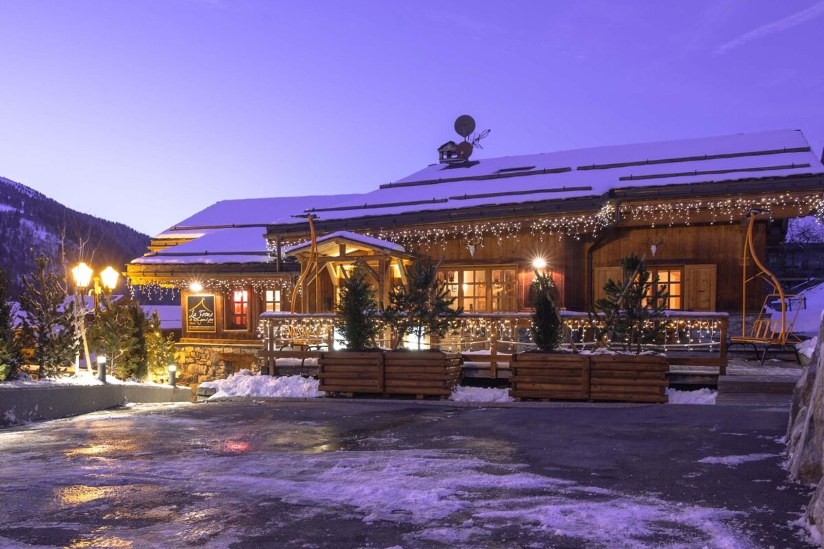A festive wooden chalet, covered in snow and twinkling with warm lights, glows under a purple twilight sky in a wintry mountain setting.