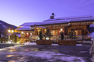 A cozy, snow-covered wooden chalet glows with festive string lights at dusk. Snowy ground and distant mountains complete the winter scene.