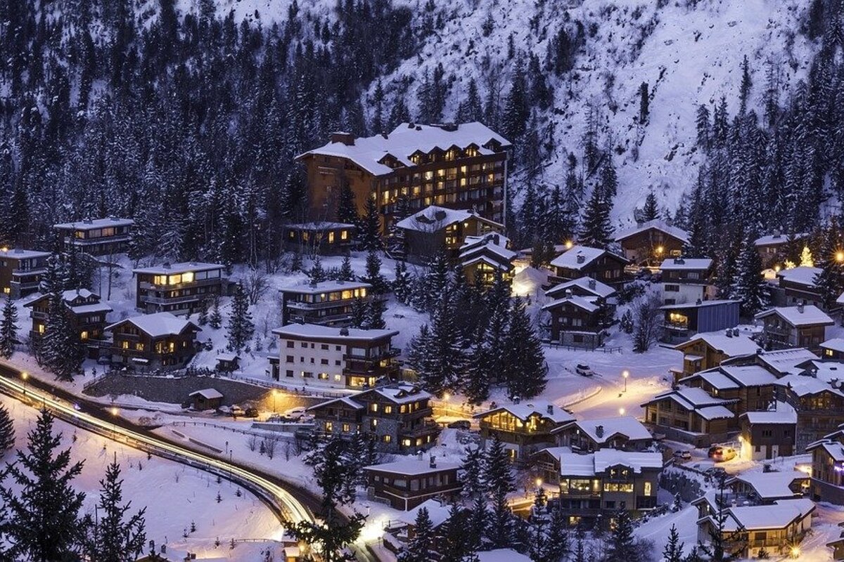 A picturesque snowy mountain village at dusk, filled with glowing wooden chalets, pine trees, and a winding road with light trails.