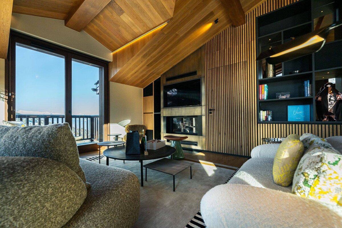 Modern chalet living room featuring warm wood-paneled ceilings, cozy sofas, a fireplace, and sliding doors opening to a scenic mountain view.