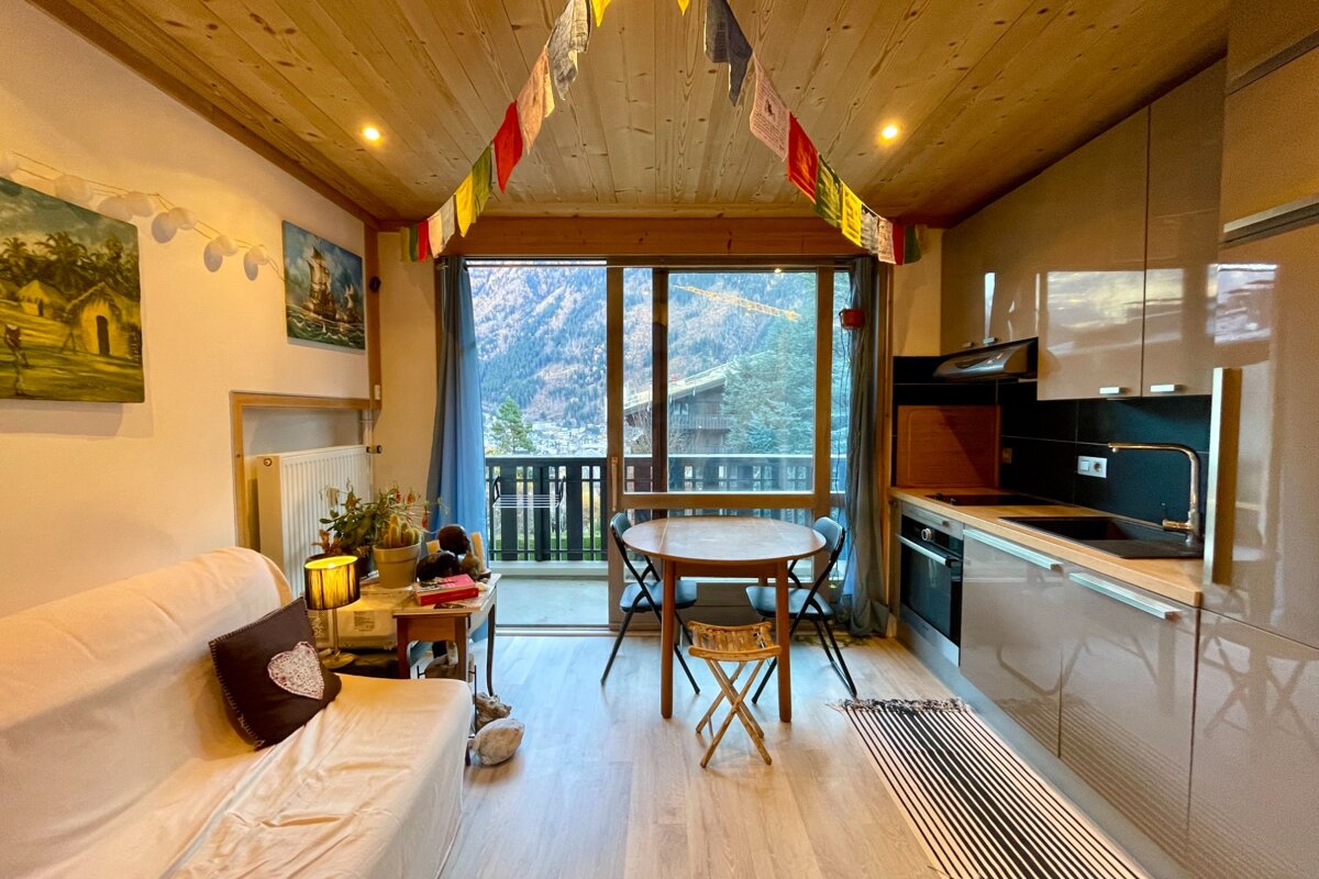 Cozy apartment with wood ceiling, mountain view balcony, kitchenette, sofa, and dining area. Prayer flags add a touch of color.