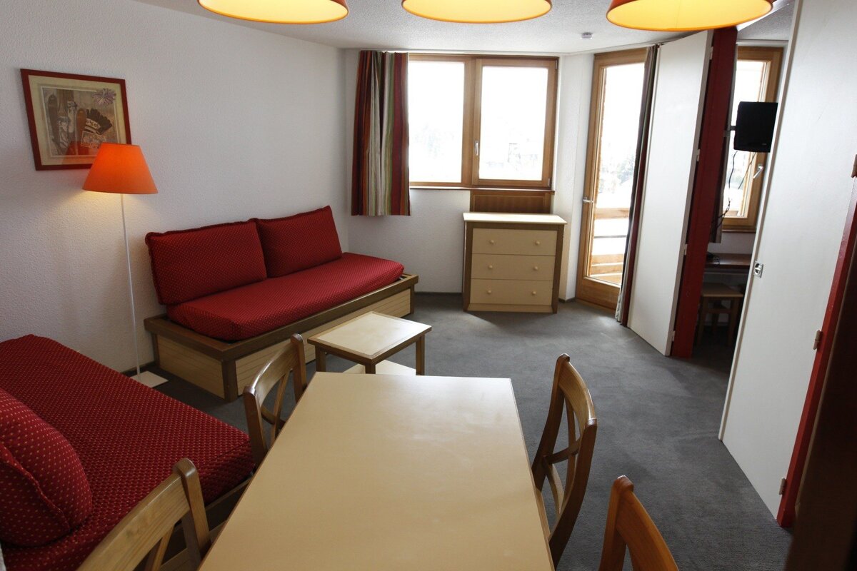 A practical living/dining room with red sofa beds, a wooden table & chairs, a dresser, an orange lamp, and large windows with striped curtains.