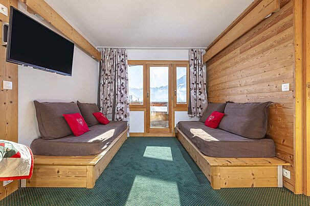 Cozy room with two brown daybeds, red pillows, green carpet, wooden walls, a TV, and a balcony door showing snowy mountains.