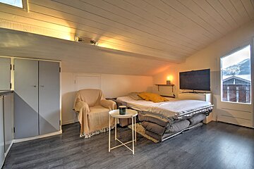 Cozy attic room with a sofa bed, armchair, TV, and a window offering a mountain view. Features a sloped ceiling and dark flooring.