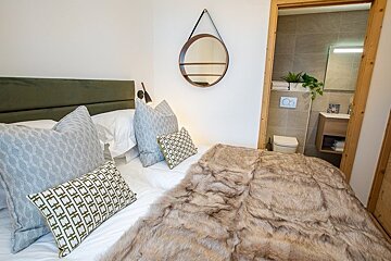 Cozy bedroom with a faux-fur blanket, decorative pillows, and a round wall mirror, looking into an adjacent bathroom.