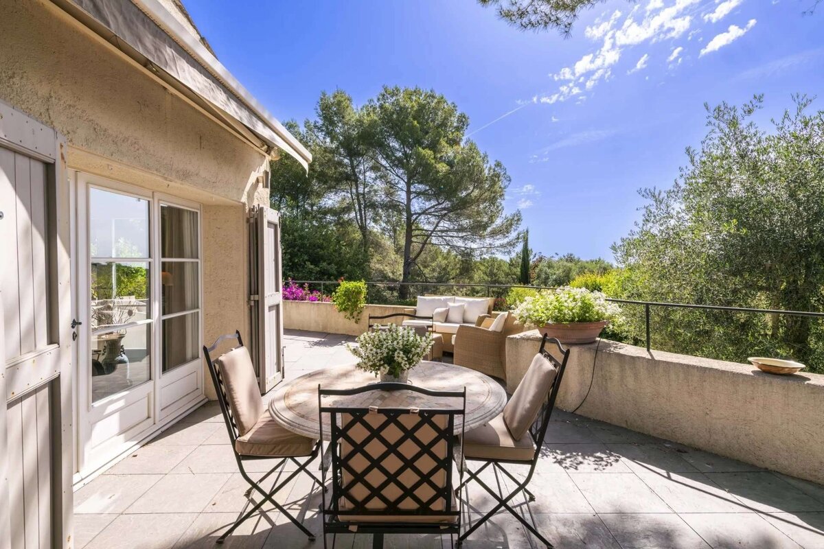 Sunny Mediterranean terrace with a dining set, lounge furniture, and lush green trees under a bright blue sky.