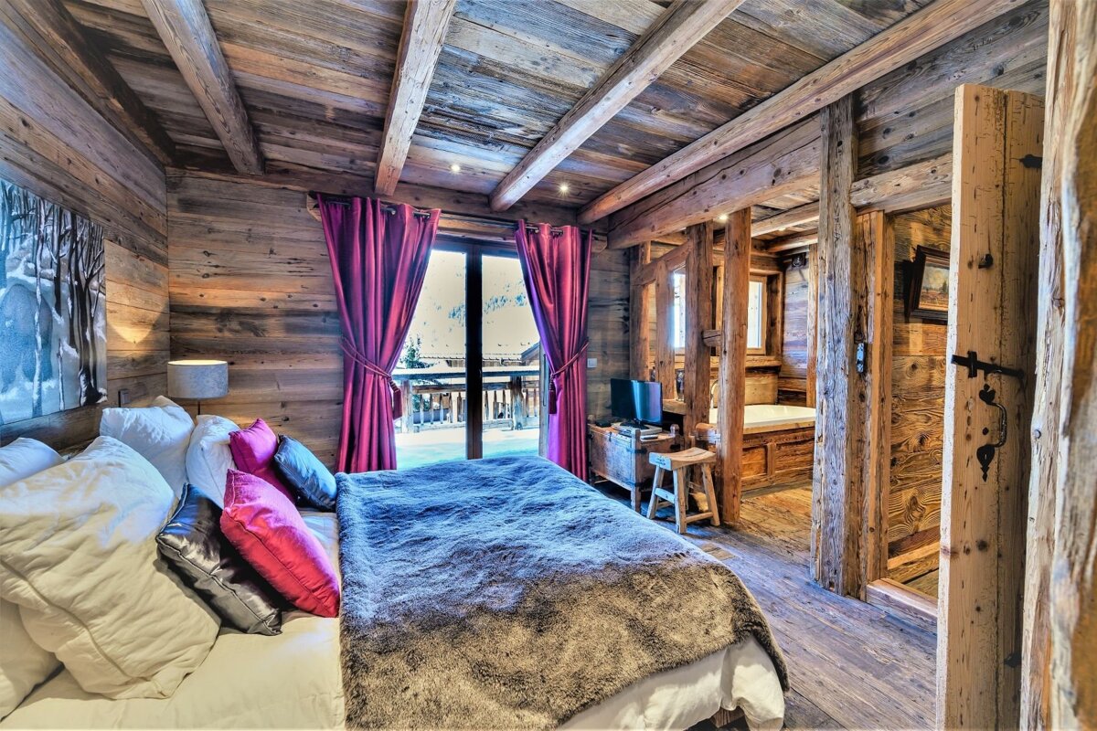 A rustic wooden chalet bedroom features a large bed with colorful pillows, red curtains, balcony view, and an open bathroom.
