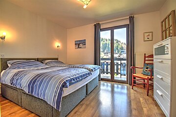 A cozy bedroom with two beds, blue striped bedding, and a balcony overlooking mountains. Features wood floors, a white dresser, and a red chair.