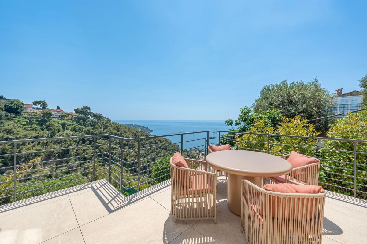 A sunny terrace with outdoor seating overlooks a breathtaking view of the sea and lush green hills.