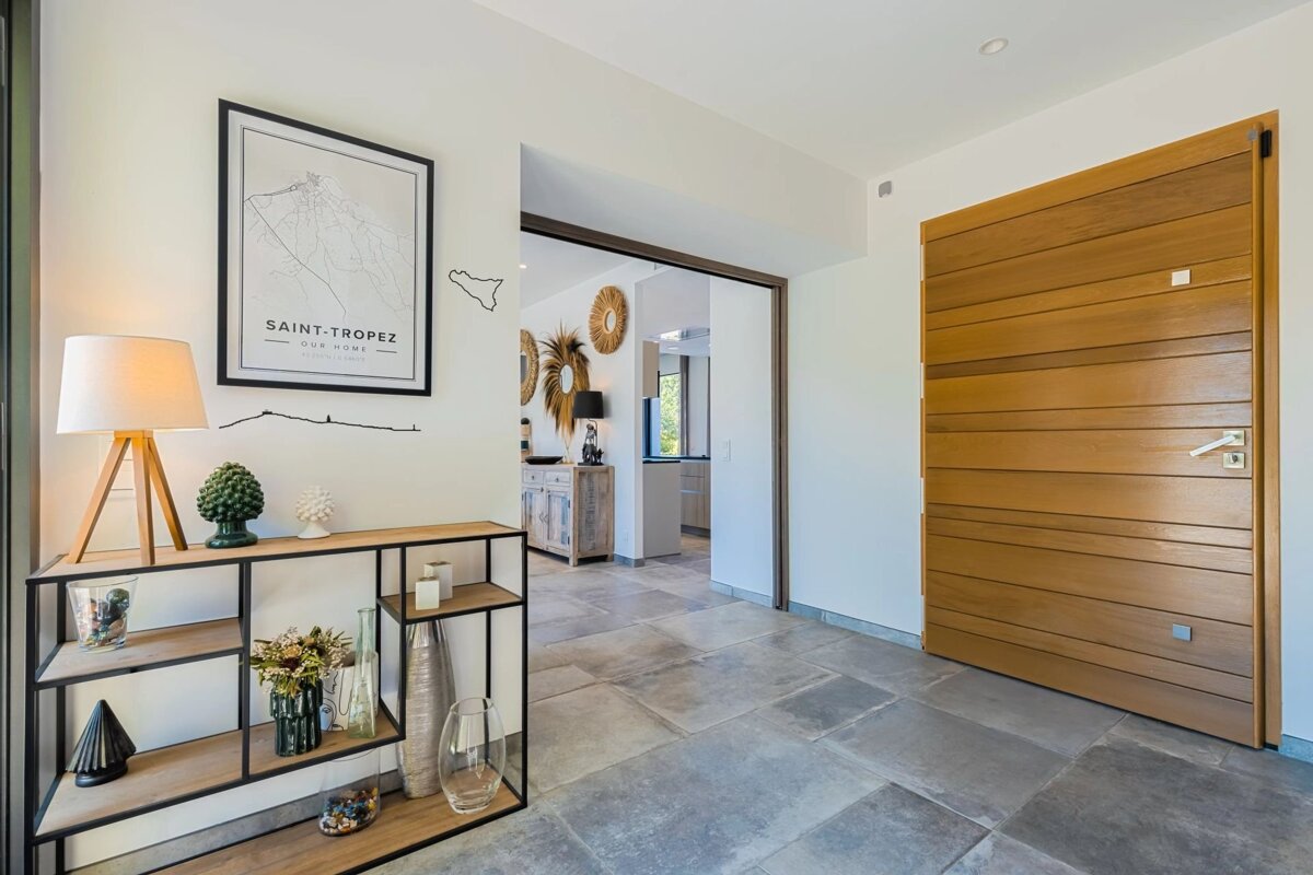 A modern home entryway with a large wooden door, stone tiled floor, and a decorated console table. An open archway leads to another room.