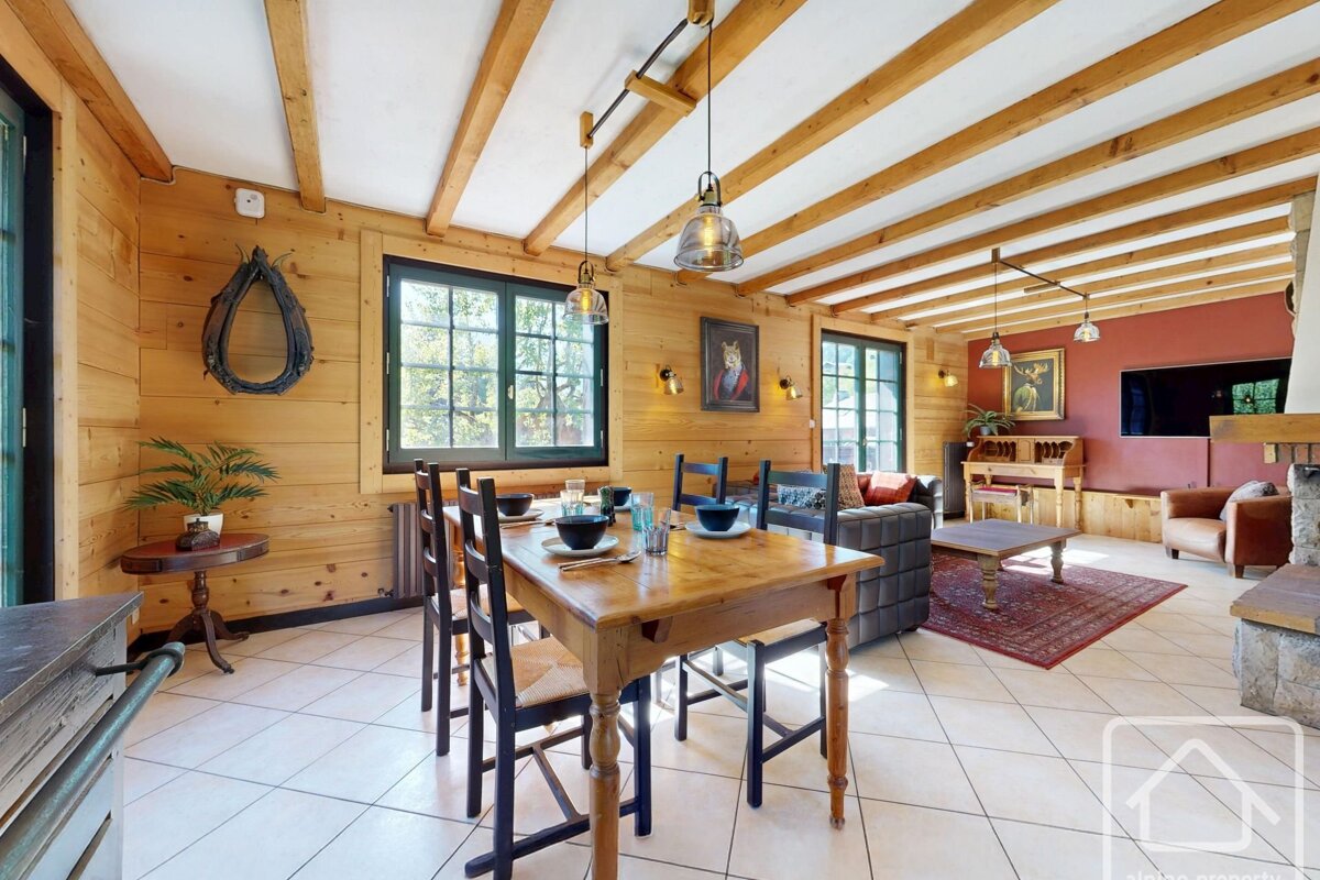A rustic wooden chalet interior with a dining table and chairs, adjacent living area, tiled floor, and exposed ceiling beams.