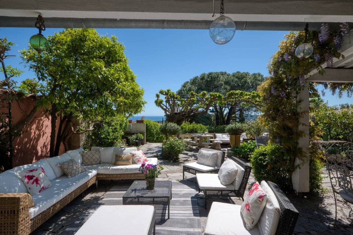 A luxurious outdoor patio with comfortable seating and lush greenery, adorned with flowers and overlooking the sea under a clear blue sky.